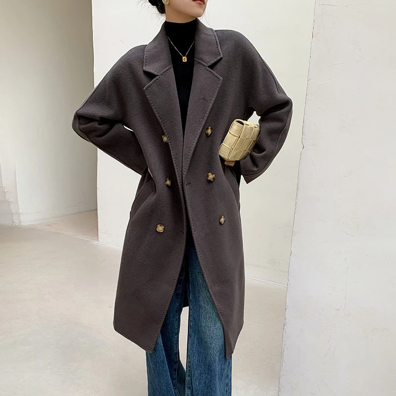 A person wearing an oversized dark brown double-breasted coat over a black turtleneck, paired with blue jeans and holding a beige quilted handbag in a minimalist indoor setting.