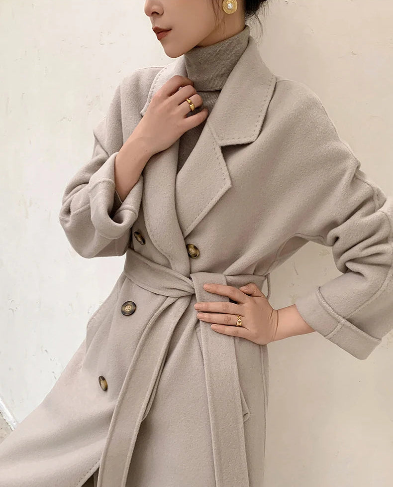 A woman wearing a light beige double-breasted cashmere coat with a matching waist tie belt, styled over a high-neck turtleneck sweater. The image features a warm, soft aesthetic with a textured off-white wall background and text overlay at the bottom reading 'WINTER CASHMERE COAT'.