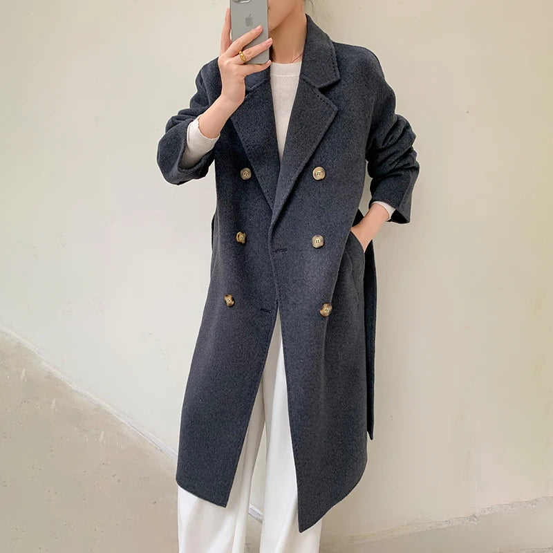 A person takes a mirror selfie wearing a dark charcoal gray double-breasted coat with prominent gold buttons, paired with a white top and white wide-leg pants, set against a plain off-white wall.