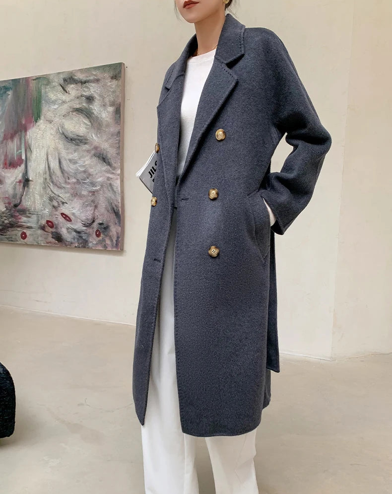 A model wears a long double-breasted cashmere coat in a dark slate gray color with gold buttons, paired with white wide-leg trousers and a white top. The image is presented as a product display with text overlays at the top and bottom.