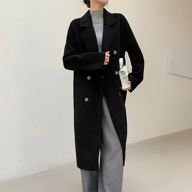 A woman wearing a long black double-breasted coat over a gray ribbed turtleneck and gray wide-leg pants, holding a white bottle in a minimalist indoor setting.