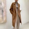 A woman models a mid-length camel brown double-breasted coat over a gray turtleneck sweater and beige wide-leg trousers, holding a white purse against a neutral indoor wall background.