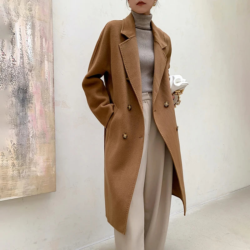 A woman models a mid-length camel brown double-breasted coat over a gray turtleneck sweater and beige wide-leg trousers, holding a white purse against a neutral indoor wall background.