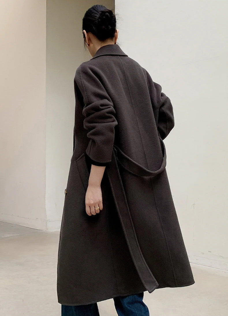 A person shown from the back wearing a long dark brown coat with a large draped waist detail, photographed in a minimalist indoor setting with off-white walls.