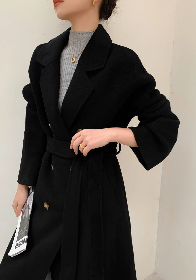 A woman wearing a black double-breasted belted coat over a gray ribbed top, holding a magazine, photographed against a plain light wall.